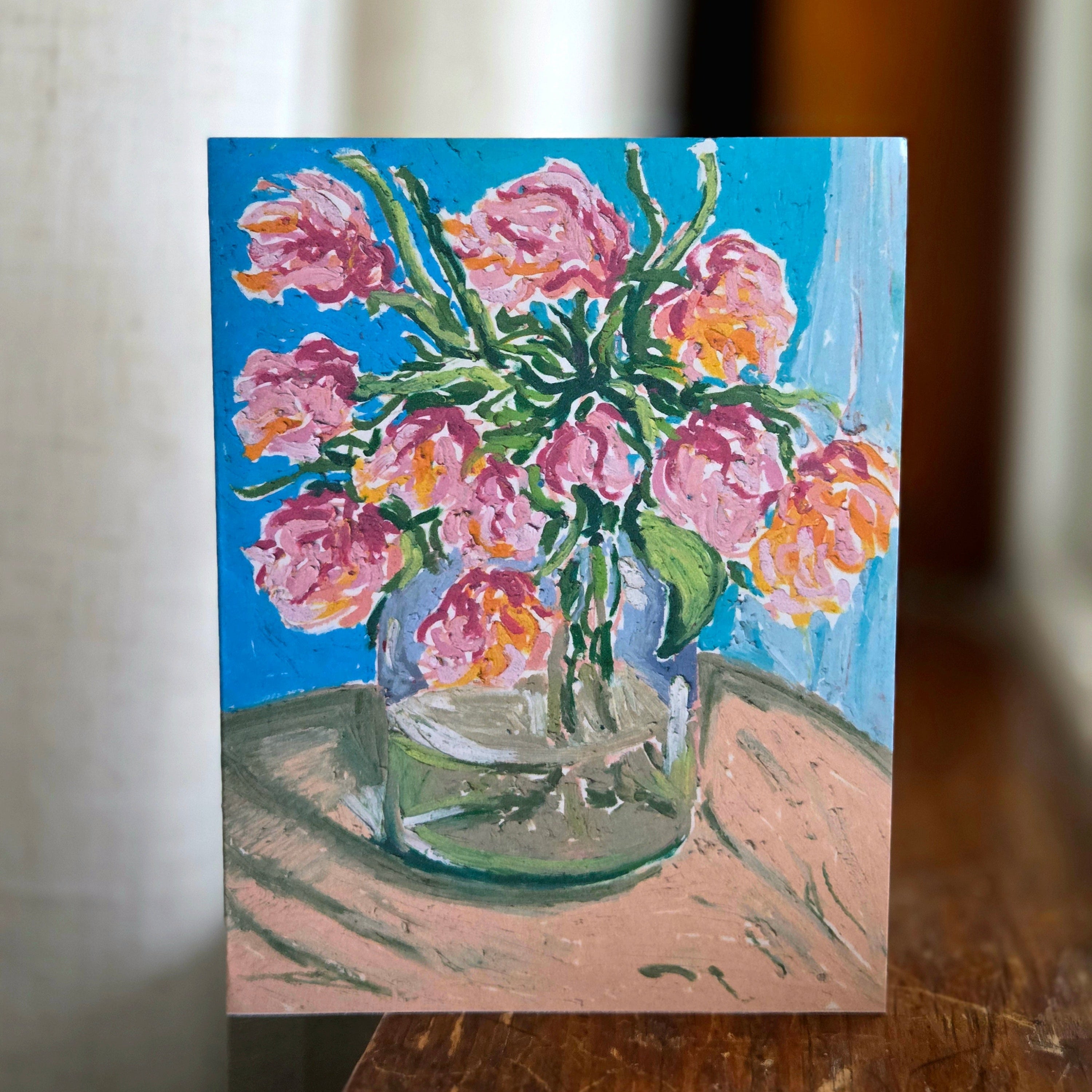 Card with a painting of pink flowers in a glass vase on a wooden surface.